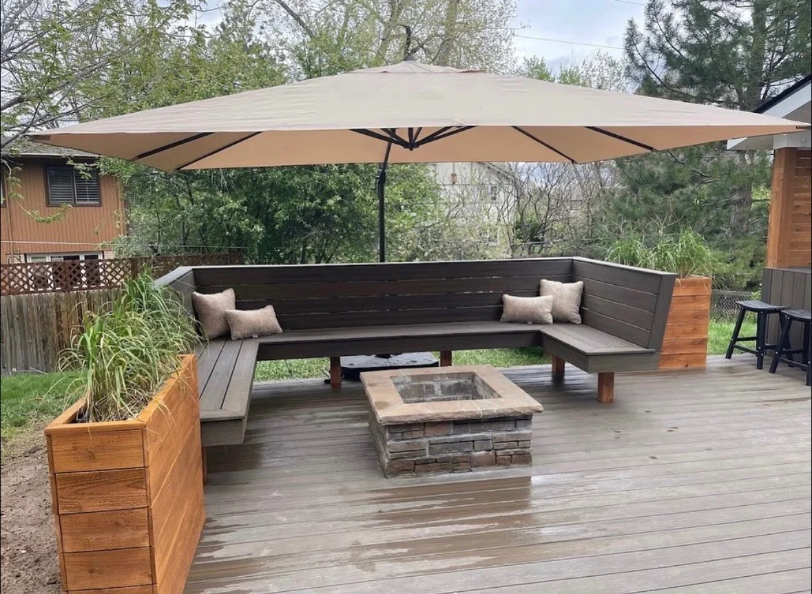 Dark composite deck with built-in bench seating, stone fire pit, and large tan umbrella