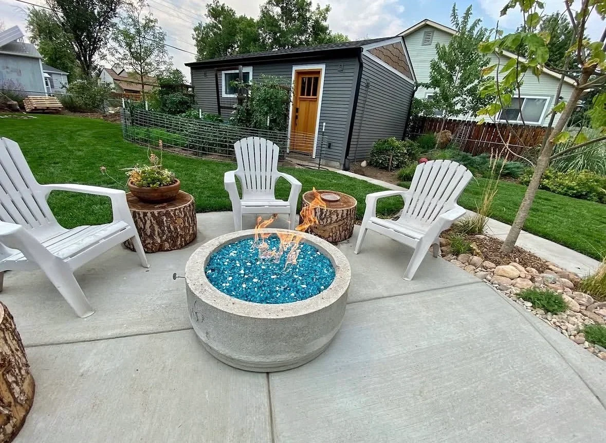 Concrete patio with circular fire pit, blue glass media, white chairs, and stone edging