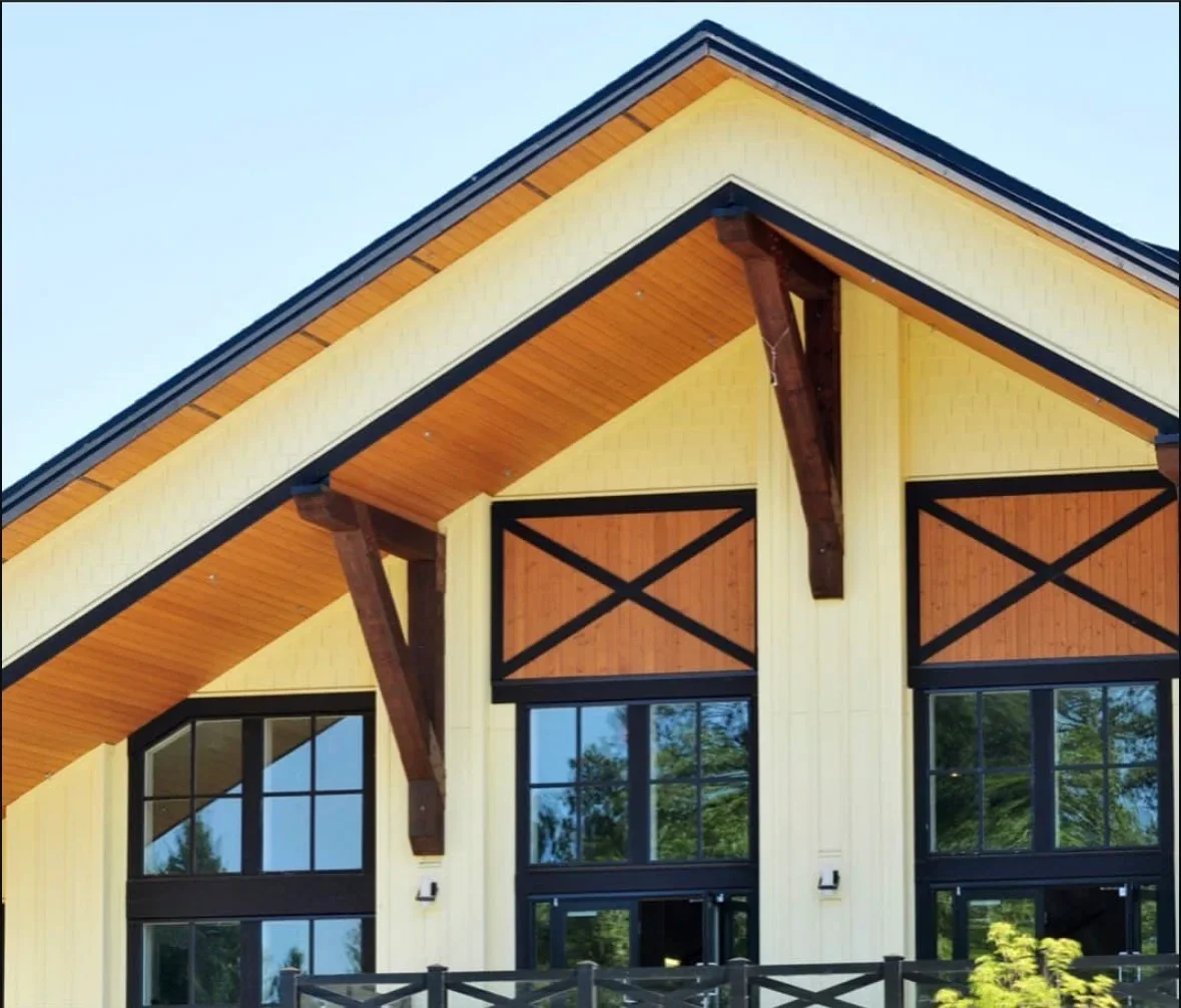 Cream siding with cedar soffit, exposed wood beams, and black-framed windows with X-shutters